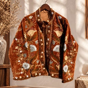 May include: A rust-colored velvet jacket featuring intricate floral embroidery in shades of cream, light blue, and dark green. The garment has a high collar, long sleeves, and a front opening, with detailed stitching along the edges and placket.