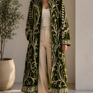 May include: A long, olive green velvet coat featuring intricate cream-colored embroidery in a swirling vine and leaf pattern. The coat has a high collar, long sleeves, and an open front, worn over a plain white top and cream-colored trousers.