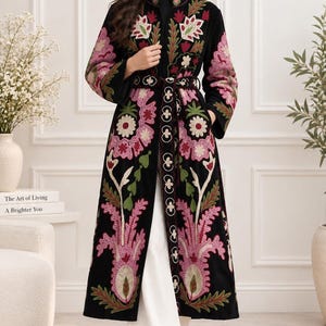 May include: A long black coat featuring intricate floral embroidery in shades of pink, green, and white. The garment has a high collar, long sleeves, and a matching tie belt. The text TASSUHANDICRAFT HANDMADE FASHION FOR A BRIGHTER YOU is visible.