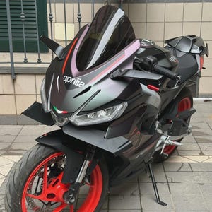 Puede incluir: Una motocicleta negra y plateada con una franja roja y blanca en el carenado delantero. La motocicleta tiene un parabrisas tintado oscuro y un asiento negro. La motocicleta está aparcada en una acera.