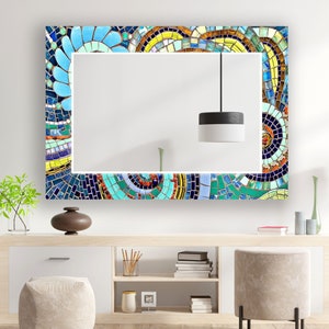 Può includere: Uno specchio rettangolare con una cornice a mosaico colorata. La cornice presenta un motivo a mosaico blu, verde, giallo e arancione. Lo specchio è appeso a una parete bianca sopra uno scaffale in legno.