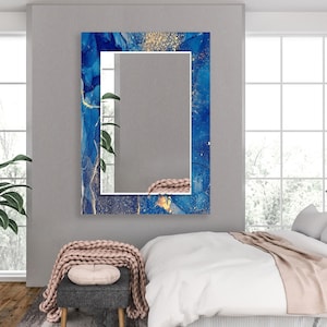 Puede incluir: Espejo de pared rectangular con un vibrante diseño abstracto azul y dorado. El espejo tiene un borde blanco y está montado en una pared gris. El diseño presenta patrones azules y dorados arremolinados, creando una estética moderna.