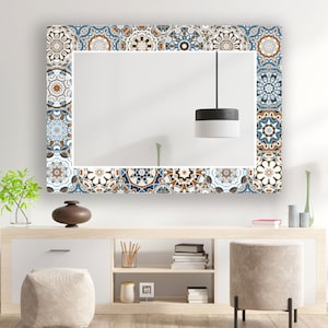 May include: A rectangular mirror with a decorative frame made of blue, brown and white patterned tiles. The mirror is hanging on a white wall above a wooden shelf with books and decorative items.