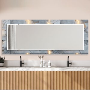 Può includere: Un mobile da bagno con due lavandini e uno specchio rettangolare grande con una cornice a motivo di marmo. Lo specchio ha un bordo bianco ed è illuminato da luci. Il mobile è in legno con una finitura marrone chiaro e ha un piano di lavoro bianco.