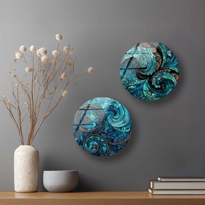 two round wall art pieces with intricate swirling designs in shades of blue and green, hanging on a gray wall above a wooden shelf. The shelf also holds a white vase with dried flowers, a gray bowl, and a stack of books.
