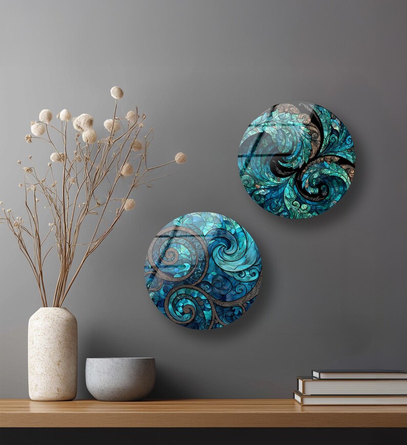 two round wall art pieces with intricate swirling designs in shades of blue and green, hanging on a gray wall above a wooden shelf. The shelf also holds a white vase with dried flowers, a gray bowl, and a stack of books.