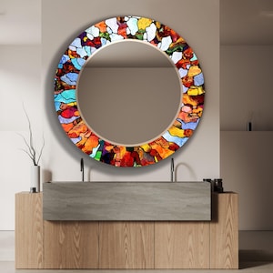 Puede incluir: Un espejo redondo con un marco de vidrio de colores. El marco presenta una variedad de colores, incluyendo rojo, naranja, amarillo, verde, azul y morado. El espejo está colgado en una pared sobre un tocador de madera con dos lavabos.
