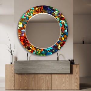 Könnte beinhalten: Ein runder Spiegel mit einem bunten Buntglasdesign. Der Spiegel ist in einem dünnen, goldfarbenen Metallrahmen gefasst. Der Spiegel hängt an einer Wand über einem Holzwaschtisch mit einer grauen Arbeitsplatte.