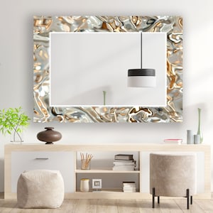 May include: A large rectangular mirror with a decorative, rippled metallic frame in shades of silver, gold, and bronze. The mirror hangs above a light-colored wooden cabinet with a modern design. A black and white pendant light hangs in front of the mirror.