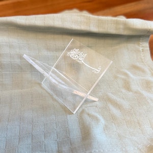 May include: A clear acrylic stand with Arabic calligraphy etched into the surface. The stand is designed to hold a book or other object.