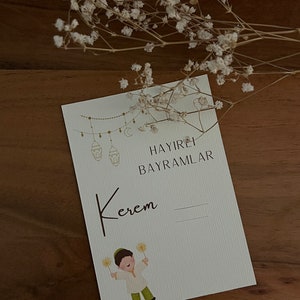 以下が含まれることがあります： 金色の三日月と提灯が紐で吊り下げられた白いカード。 「HAYIRLI BAYRAMLAR」というテキストが黒で印刷されています。 「Kerem」という名前が筆記体で書かれています。 テキストの下には、火花を持った少年の漫画イラストがあります。