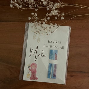 以下が含まれることがあります： 「Hayirli Bayramlar」というテキストと、筆記体で「Melis」という名前が書かれた白いカード。カードには、ピンクの頭巾をかぶった女の子が提灯を持っている水彩画が描かれている。カードには、青と白の模様のギフトカードが2枚付いています。