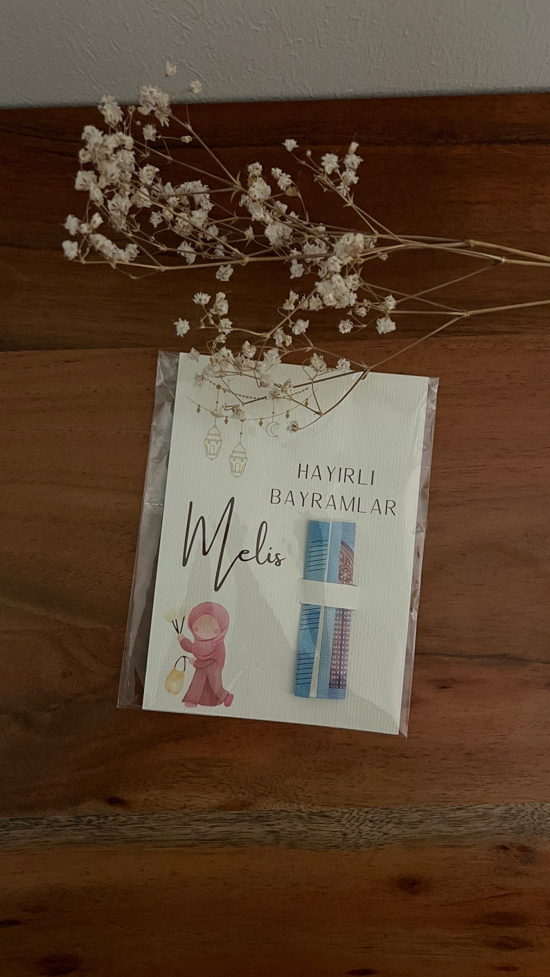 以下が含まれることがあります： 「Hayirli Bayramlar」というテキストと、筆記体で「Melis」という名前が書かれた白いカード。カードには、ピンクの頭巾をかぶった女の子が提灯を持っている水彩画が描かれている。カードには、青と白の模様のギフトカードが2枚付いています。