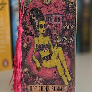 May include: A rectangular wooden bookmark featuring a green-skinned figure in a skull-patterned bikini sitting on a pink flamingo pool float. The illustration includes a mansion, palm trees, and the text "HOT GHOUL SUMMER" with a pink tassel.