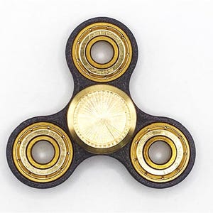 May include: A black and gold fidget spinner with a three-lobed design. The spinner has three circular bearings with gold-coloured metal rims and a central gold-coloured disc. The body is black.