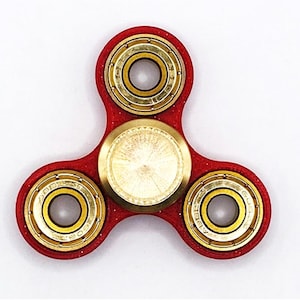 May include: A red fidget spinner with three arms and gold-coloured bearings. The centre and bearing rings have a metallic finish. The design is symmetrical, with a circular bearing at each end of the arms.