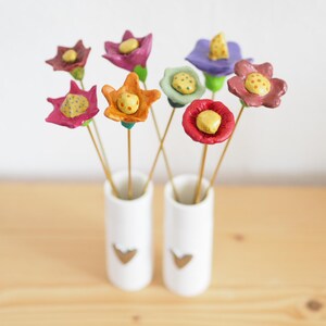 Könnte beinhalten: Eine Sammlung handgefertigter Tonblumen in einer weißen Vase mit goldenem Herzdesign. Die Blumen haben verschiedene Farben, darunter Rosa, Orange, Lila und Rot, mit gelben Zentren. Die Vase steht auf einer hellen Holzoberfläche.