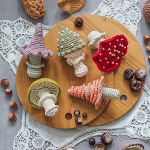 May include: A collection of six crocheted mushroom ornaments in various colours including lavender, sage green, red with white spots, yellow-green, and peach. The mushrooms feature textured caps and stems, arranged on a round wooden board with scattered hazelnuts.