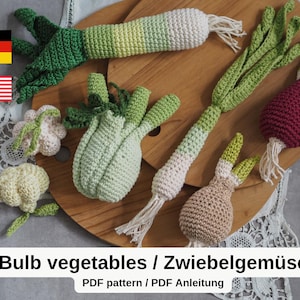 May include: A selection of crocheted bulb vegetables, such as leeks, onions, and radishes, displayed on a wooden board. The vegetables are in green, white, and red. The image includes the text "Bulb vegetables / Zwiebelgemüse".
