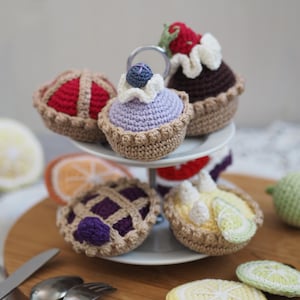 Può includere: Una collezione di mini torte e crostate all'uncinetto in vari colori, tra cui rosso, viola e marrone. I dessert sono disposti su un supporto a più livelli bianco e una tavola di legno, con posate.