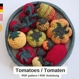May include: A selection of crocheted tomatoes in shades of red, orange, yellow, and green, displayed on a circular, grey-blue platter. The tomatoes vary in size and shape, with dark green or grey crocheted stems. The text "Tomatoes / Tomaten" is at the bottom.