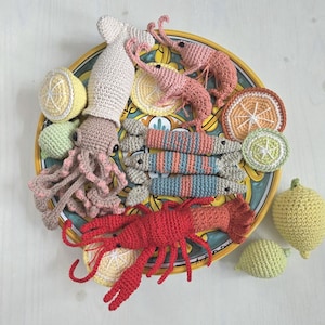 May include: A crocheted seafood platter featuring a squid, shrimp, and a lobster in shades of pink, red, and beige. The platter includes crocheted lemon and lime slices, and whole lemons. The plate has a yellow and blue floral design.