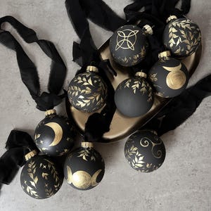 May include: A collection of black Christmas ornaments with gold leaf and celestial designs. Each ornament has a gold cap and is tied with a black ribbon. The ornaments are displayed on a gold tray.