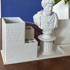 Op de afbeelding: Een grijze bureau-organizer met een buste van een Romeinse keizer. De organizer heeft een citaat van Marcus Aurelius: "Het geluk van je leven hangt af van de kwaliteit van je gedachten."