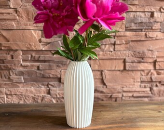 May include: A white, ribbed vase holds two vibrant pink peonies with green leaves. The vase sits on a wooden surface against a textured brick wall, creating a simple yet elegant floral arrangement.