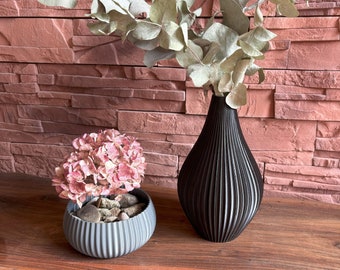 May include: A black vase with a ribbed design holds dried eucalyptus branches. A small gray planter with a ribbed design holds pink hydrangeas and small stones. The vase and planter are on a wooden surface.