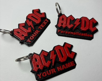 May include: Three black and red 3D printed keychains with the AC/DC logo. The keychains have the text "Your Text", "Personalization", and "Your Name" on them.