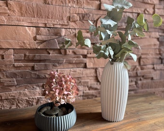 May include: A white ceramic vase with vertical ridges holds a bouquet of green eucalyptus branches. A small black planter with a similar ribbed design holds a dried pink flower arrangement.