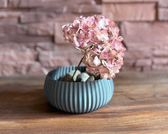 May include: A blue ceramic planter with a ribbed design, filled with small stones and a dried pink hydrangea flower.