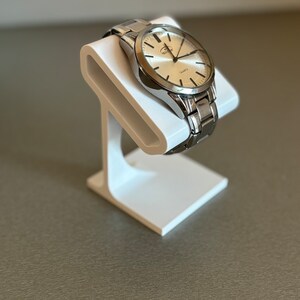 Puede incluir: Un reloj plateado con una esfera blanca y agujas plateadas se muestra en un soporte de reloj blanco. La esfera del reloj tiene las palabras "Quartz" y "Watling" impresas en ella.