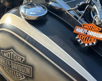 May include: Close-up of a black motorcycle gas tank with a silver stripe and a Harley-Davidson logo. A Harley-Davidson keychain with the words "Motor Harley-Davidson Cycles Fat Boy" is attached to the tank.