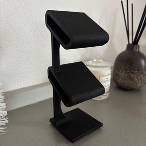 Puede incluir: Un soporte negro de dos niveles con una base cuadrada. El soporte est&aacute; dise&ntilde;ado para sostener objetos peque&ntilde;os, como joyas o relojes.