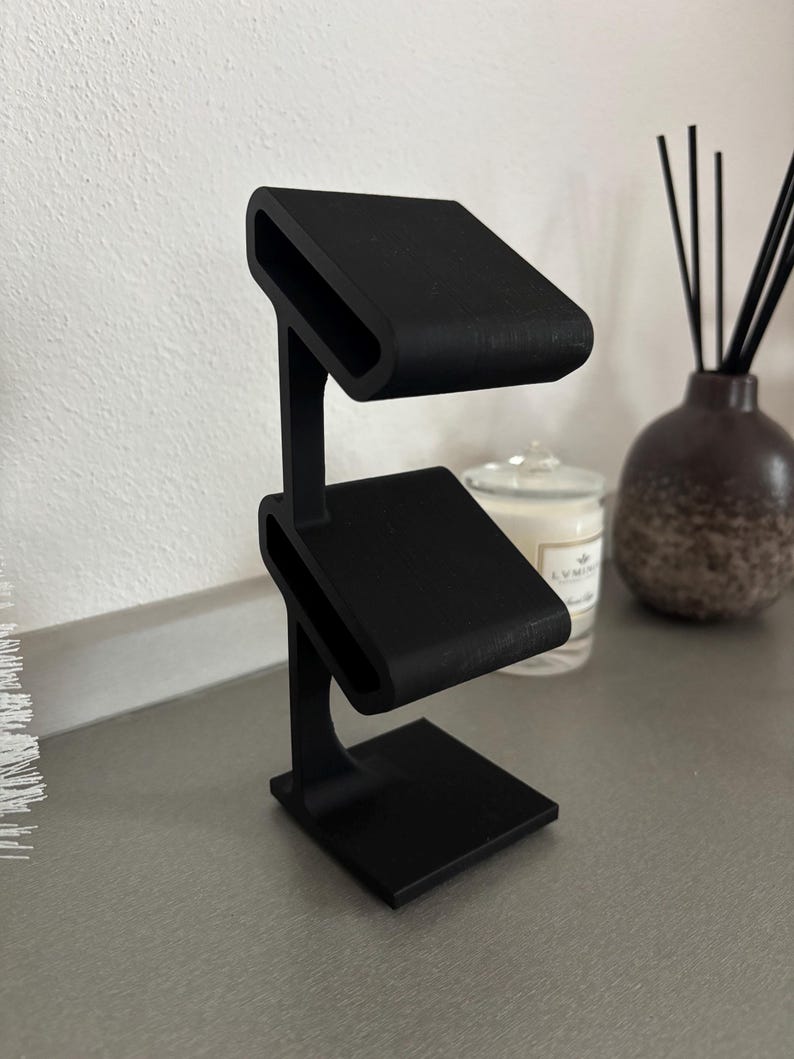 Puede incluir: Un soporte negro de dos niveles con una base cuadrada. El soporte est&aacute; dise&ntilde;ado para sostener objetos peque&ntilde;os, como joyas o relojes.