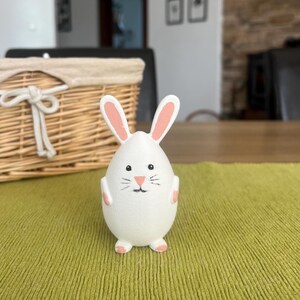 Può includere: Statuetta di coniglietto pasquale bianco con interno orecchie, naso e piedi rosa. Il coniglietto ha bocca e baffi neri dipinti. Il corpo a forma di uovo poggia su una superficie verde, con un cestino di vimini sullo sfondo.
