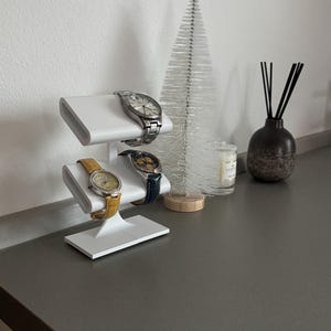 Puede incluir: Un soporte para relojes blanco de dos niveles, que sostiene tres relojes de pulsera con correas de diferentes colores. El soporte est&aacute; sobre una encimera gris con un peque&ntilde;o &aacute;rbol de Navidad artificial blanco y una botella difusora negra y marr&oacute;n.