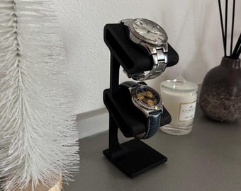 May include: A black watch display stand holding two wristwatches. The top watch has a silver band and face, while the bottom watch has a black leather band and gold face. The stand is on a gray surface.