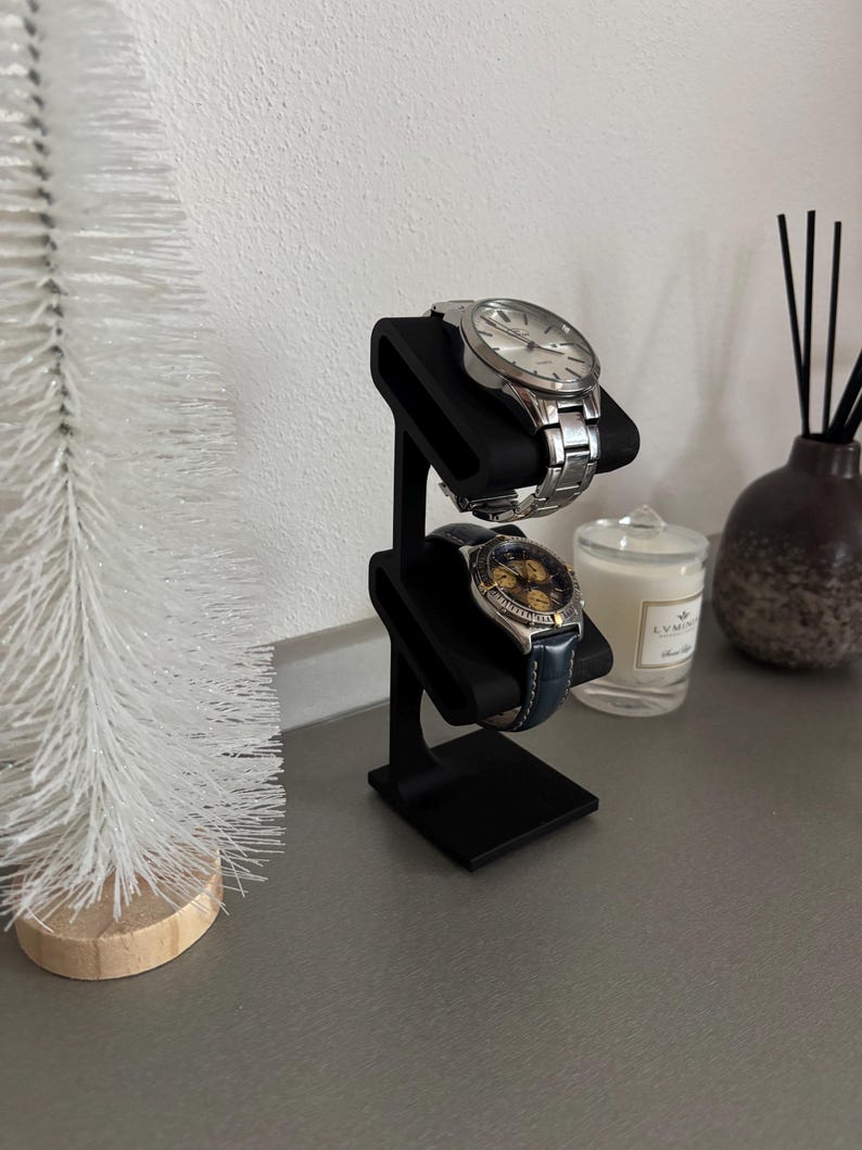 Puede incluir: Un soporte para relojes negro con dos niveles, cada uno con un coj&iacute;n negro para sujetar un reloj. Dos relojes se muestran en el soporte, uno con una correa plateada y otro con una correa azul.