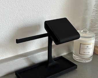May include: A matte black jewelry display stand. The stand features a rectangular base, a vertical support, and a horizontal bar. A small, rectangular platform is at the top. A candle in a glass jar is in the background.
