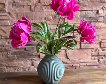 May include: A bouquet of three pink peonies in a gray, ribbed ceramic vase. The vase is sitting on a wooden table.