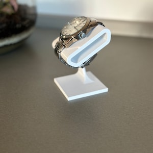 Puede incluir: Un soporte de reloj blanco con un reloj apoyado en &eacute;l. El reloj tiene una banda plateada y una esfera blanca.