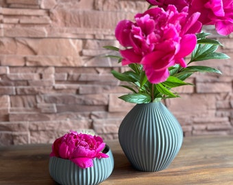 May include: Two gray ceramic vases with pink peonies. The larger vase is taller and narrower than the smaller vase. Both vases have a ribbed texture.