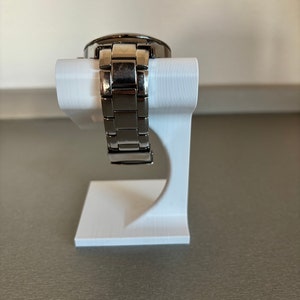 Puede incluir: Un reloj de pulsera plateado con una correa de metal se muestra en un soporte blanco. El reloj est&aacute; sobre una superficie blanca.