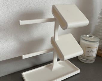 May include: A white, minimalist jewelry display stand. It features a rectangular base, a central support with two angled platforms, and two horizontal bars. A candle in a glass jar is visible in the background.