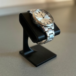 Puede incluir: Un reloj de pulsera plateado con una esfera blanca y marcas negras. El reloj est&aacute; en un soporte de reloj negro.