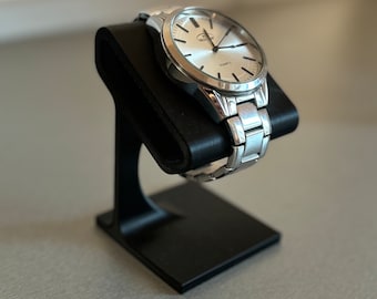 May include: A silver wristwatch with a white face and silver strap is displayed on a black watch stand. The watch has a round face with black markers and hands. The watch stand is a simple, modern design.