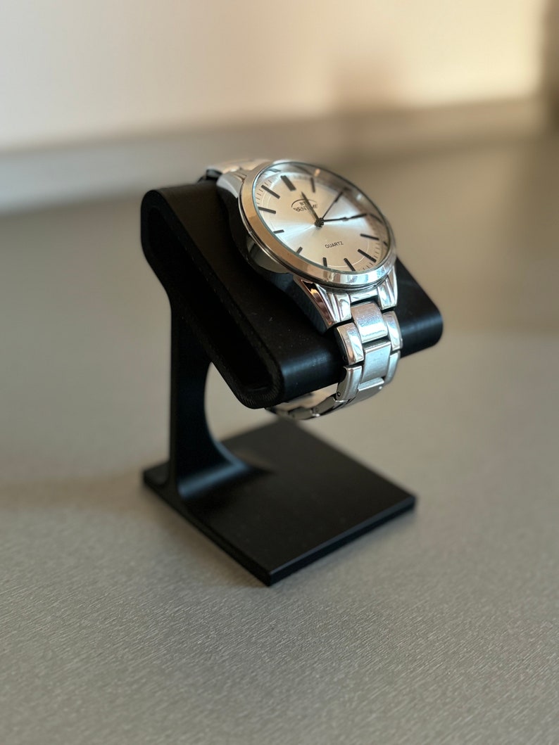 Puede incluir: Un reloj de pulsera plateado con una esfera blanca y marcas negras. El reloj est&aacute; en un soporte de reloj negro.